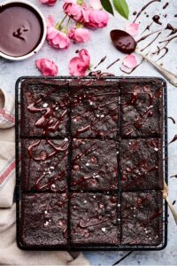 Brownies cut into 9 pieces, and set on a drying rack. There are pink flower pedals, a white cup with melted chocolate in it, and a white and red napkin next to the brownies.