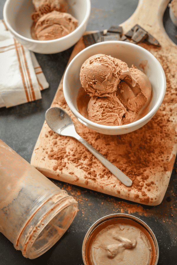 Chocolate ice cream in a white bowl. The bowl is set on top of a wooden cutting board and there's a spoon next to it.