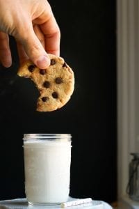 A chocolate chip cooking with a bite taken out of it, about to be dunked into a glass of milk.