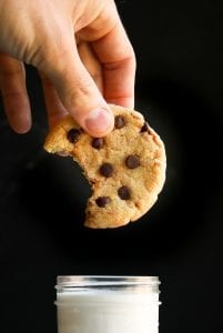 A chocolate chip cookie with a bite taken from it about to be dunked into a glass of milk.
