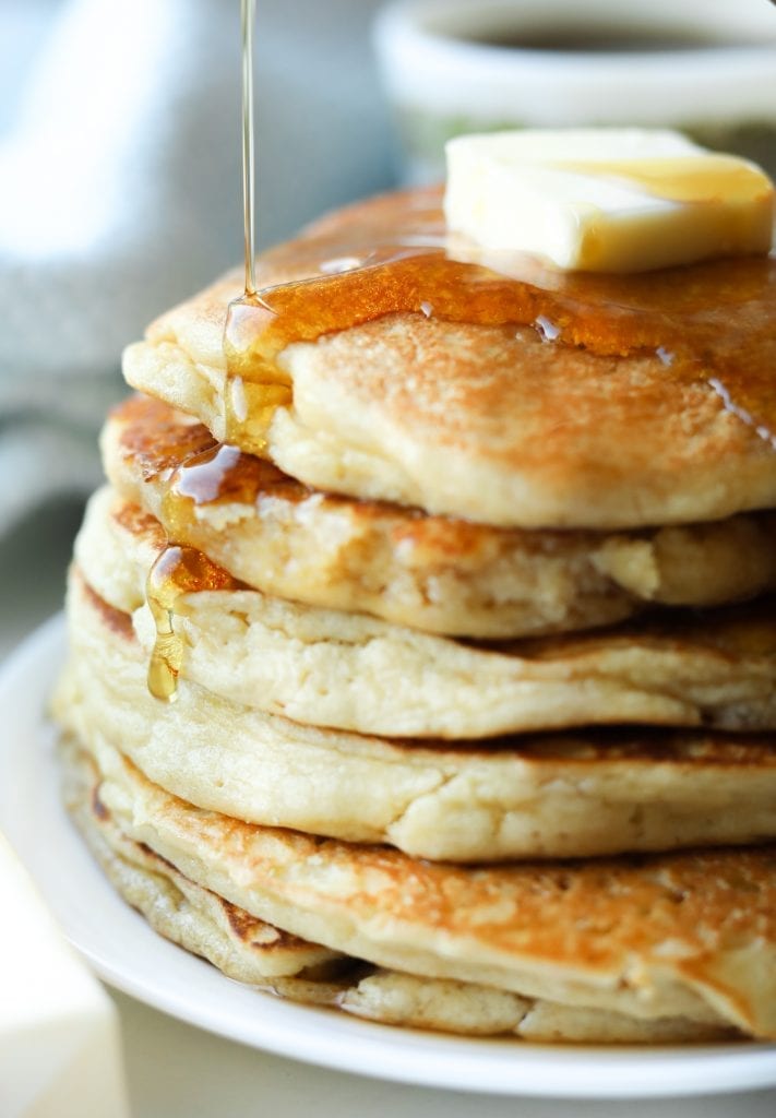 A stack of six pancakes laying on a plate with syrup running down the side of them.