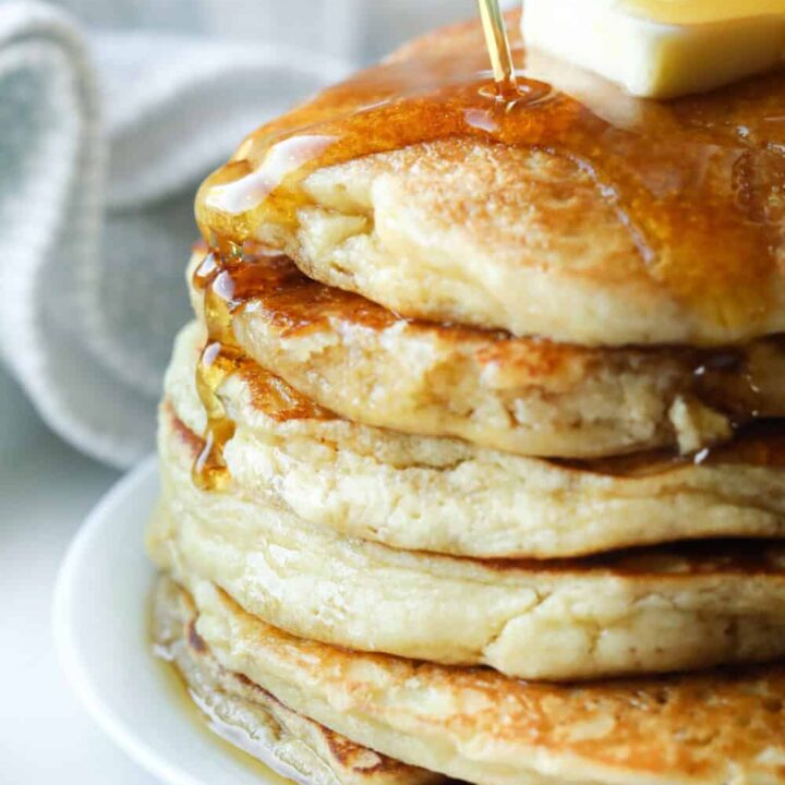 A stack of pancakes on a plate with syrup being poured on them.