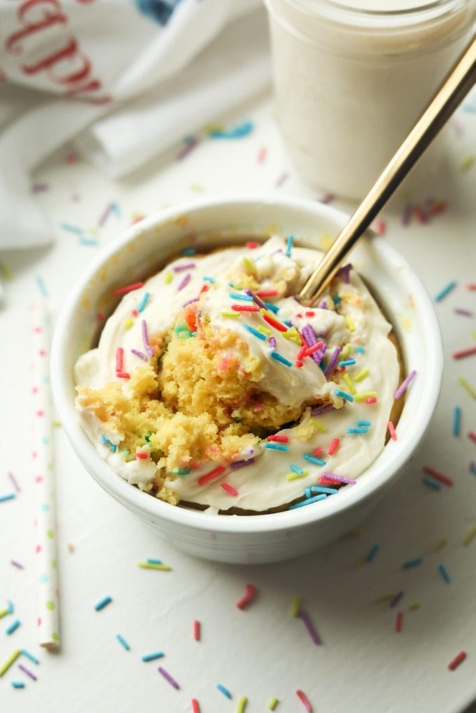 A birthday cake flavored mug cake set on a table with sprinkles surrounding it.