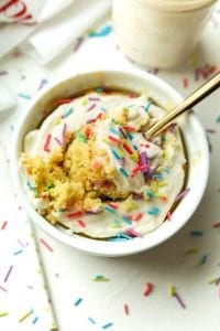 A birthday cake mug cake topped with frosting and sprinkles, and a fork holding a piece of cake.