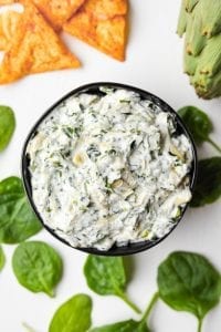 A bowl of keto spinach and artichoke dip surrounded by low carb chips, spinach, and an artichoke.