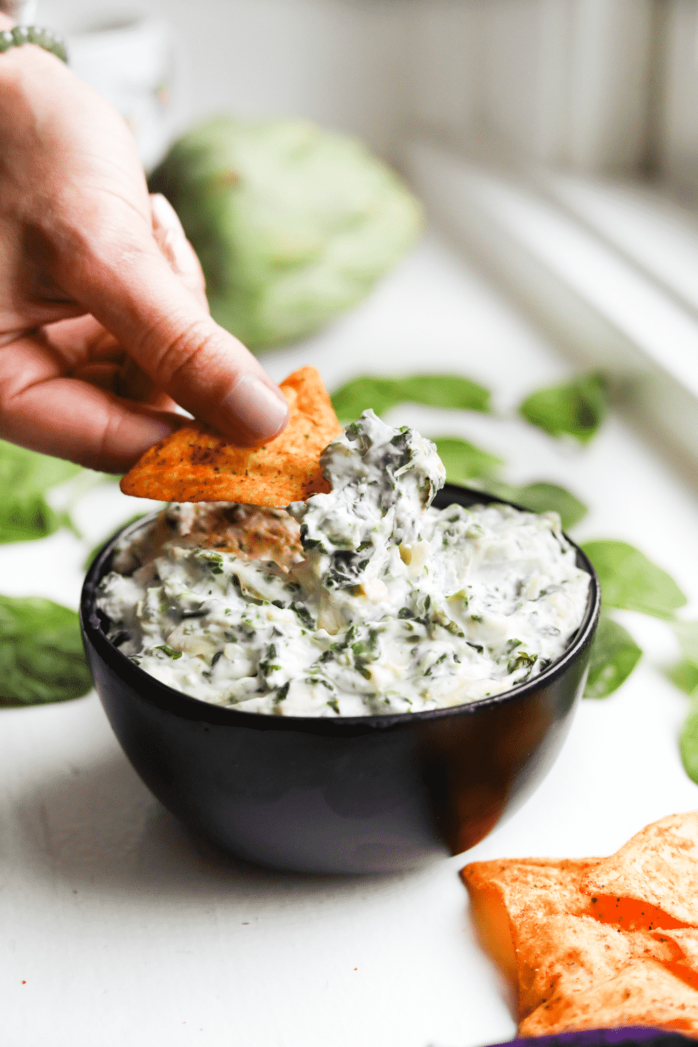 A hand holding a low carb chip with spinach and artichoke dip on it.