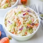 Keto coleslaw in a small white bowl, surrounded by carrots, cabbage, and a blue napkin.