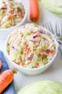 Keto coleslaw in a small white bowl, surrounded by carrots, cabbage, and a blue napkin.