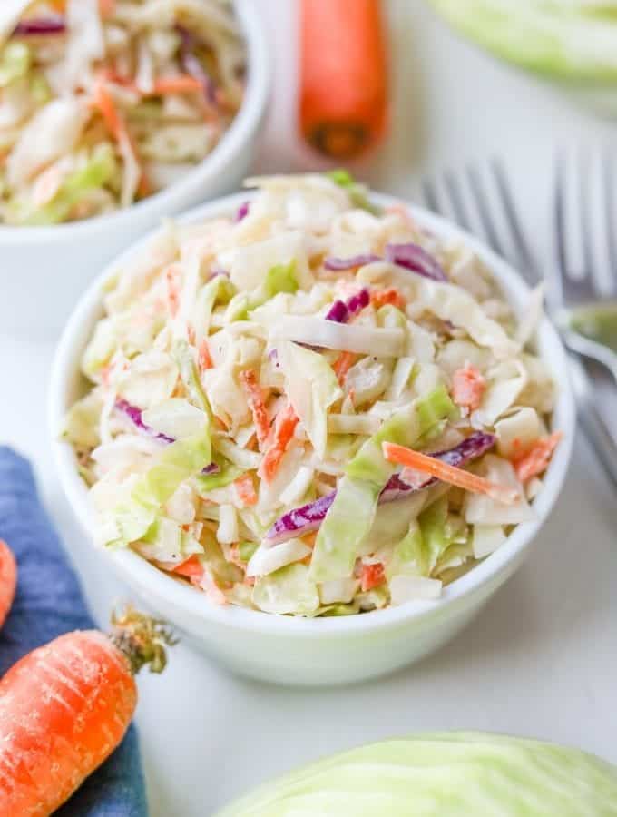 Keto coleslaw in a small white bowl, surrounded by carrots, cabbage, and a blue napkin.