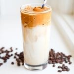 A glass filled with dalgona coffee and milk surrounded by coffee beans.