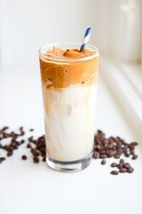 A glass filled with dalgona coffee and milk surrounded by coffee beans.