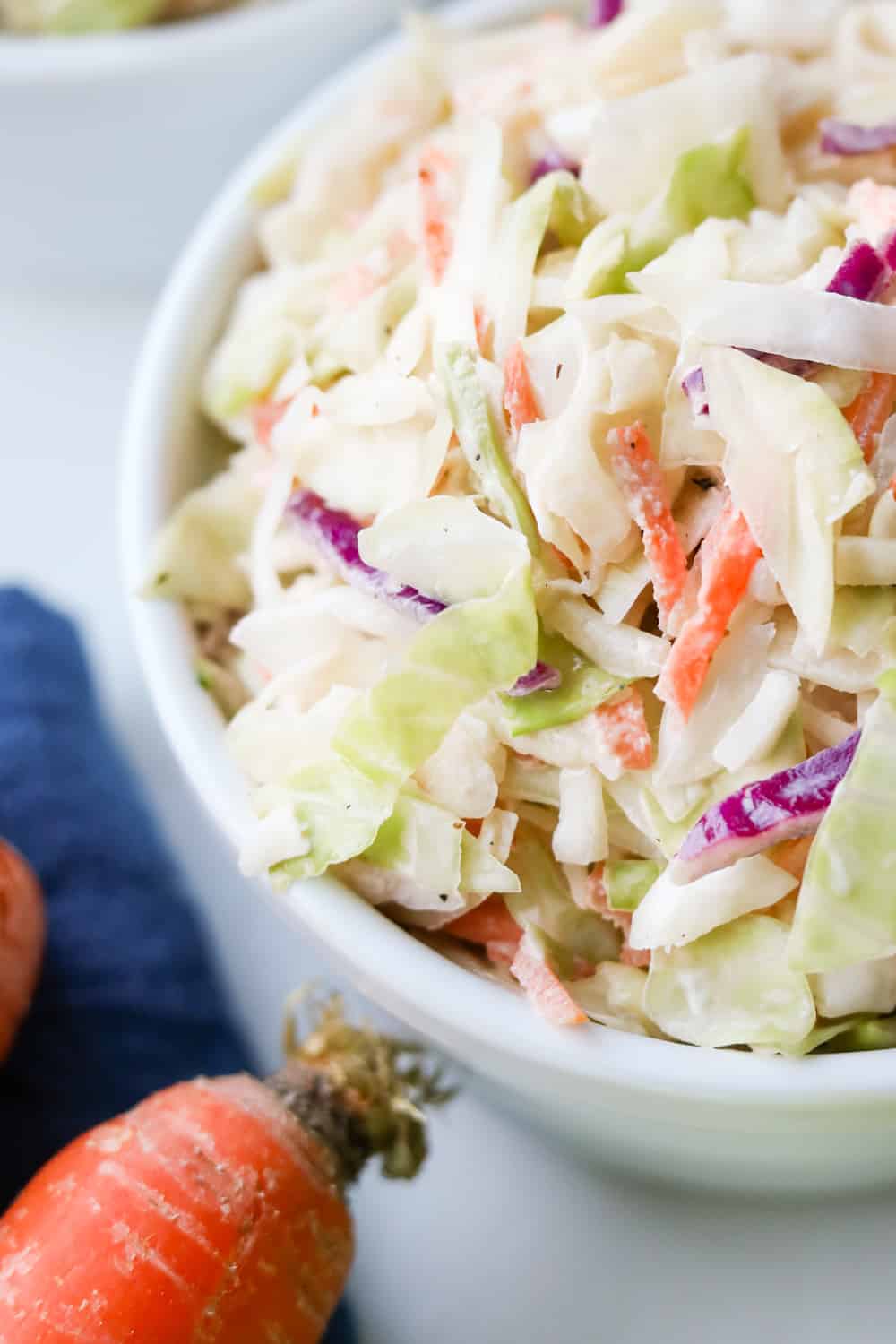 Keto coleslaw in a white bowl with a blue napkin, and carrots next to it.