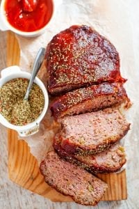 A meatloaf covered in ketchup cut into slices with spices next to it.