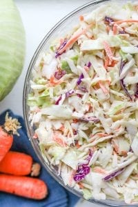 Coleslaw in a large glass bowl next to pieces of carrots and green cabbage.