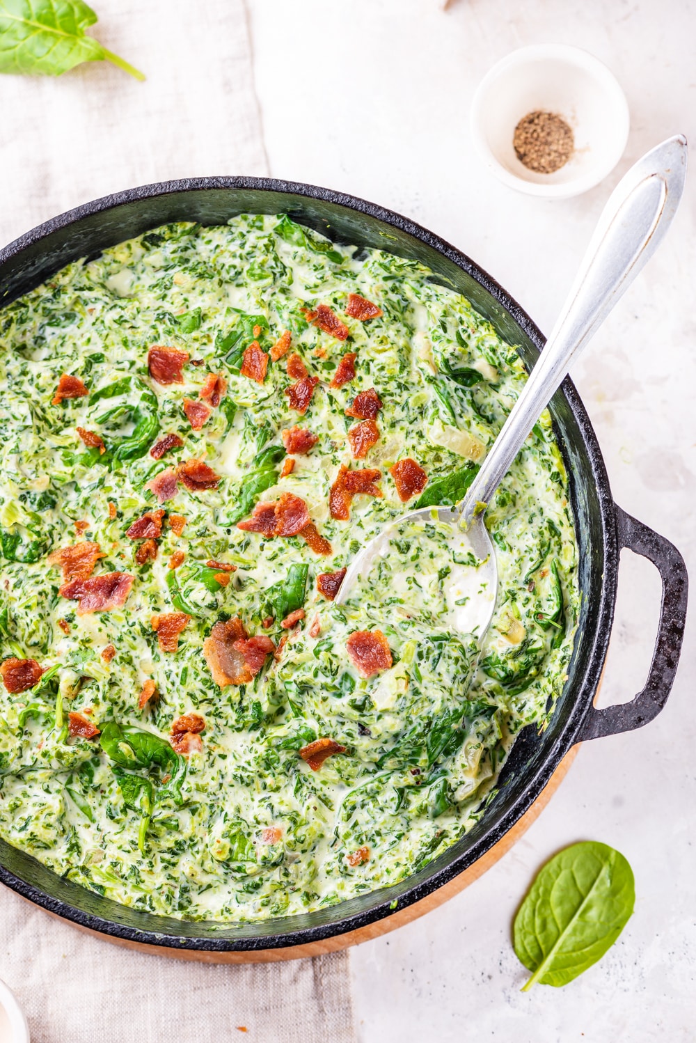 Creamed spinach in a cast iron pan surrounded by a couple of leaves of fresh spinach.