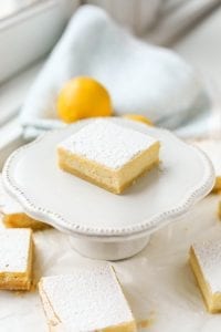 A lemon bar on a small serving dish surrounded by other lemon bars, and lemons in the background.