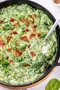 A spoon about to scoop out a portion of creamed spinach from a cast iron pan.