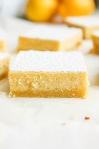 Lemon bars on parchment paper with lemons in the background.