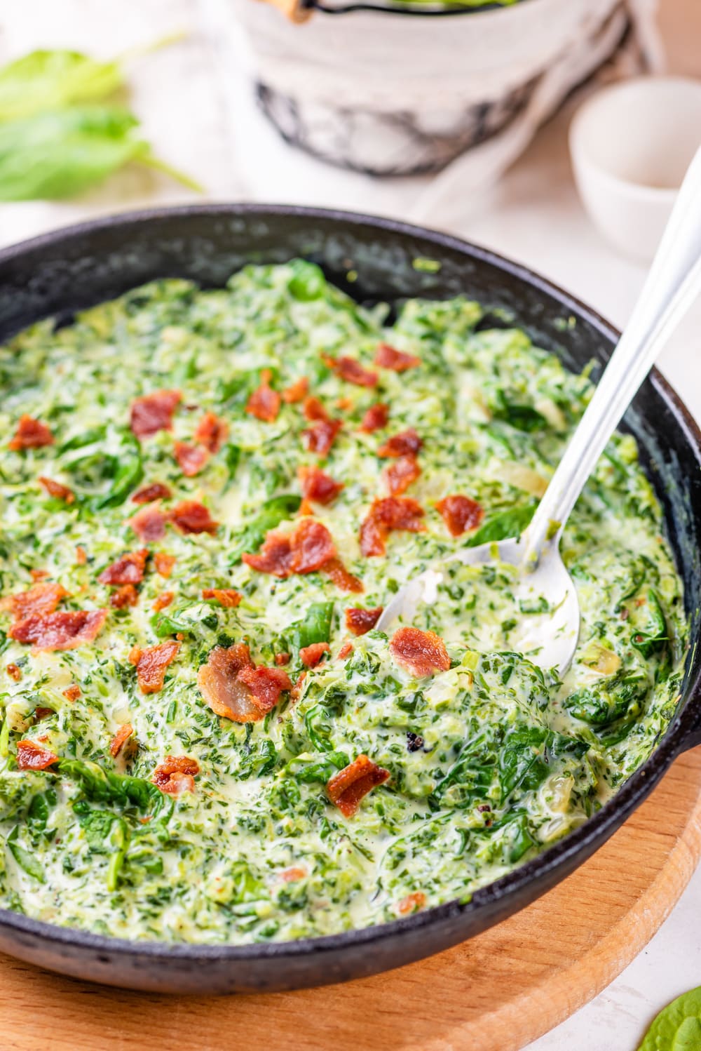 A cast iron pan filled with creamed spinach and bacon crumbles with a spoon for serving in the pan.