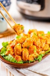 Chop sticks holding a piece of orange chicken with a bowl of orange chicken below it.