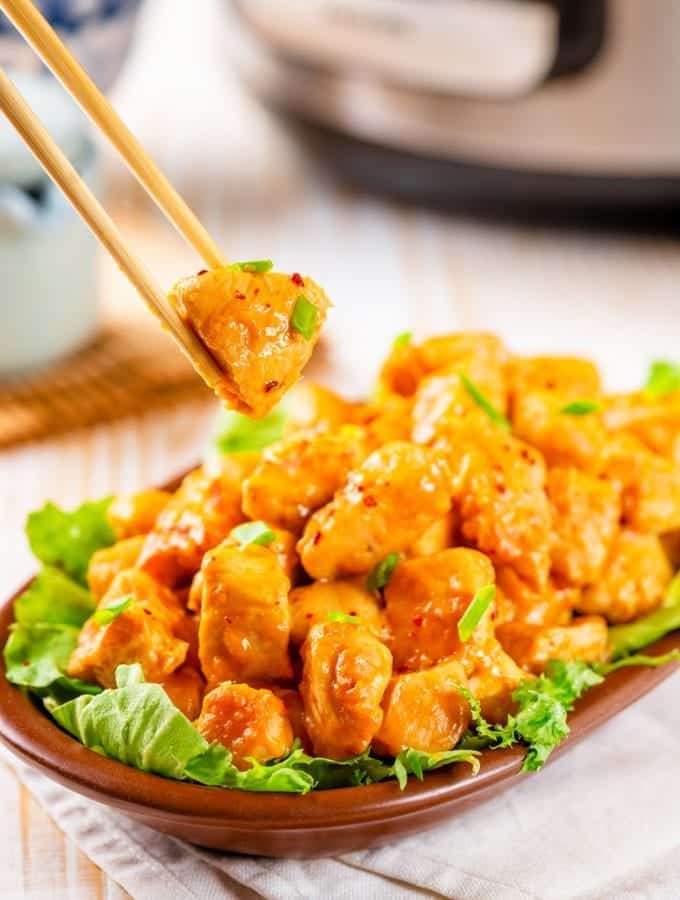 Chop sticks holding a piece of orange chicken with a bowl of orange chicken below it.