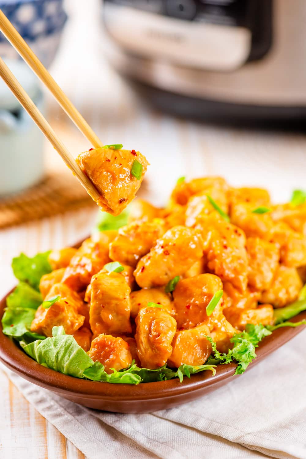 Chop sticks holding a piece of orange chicken with a bowl of orange chicken below it.