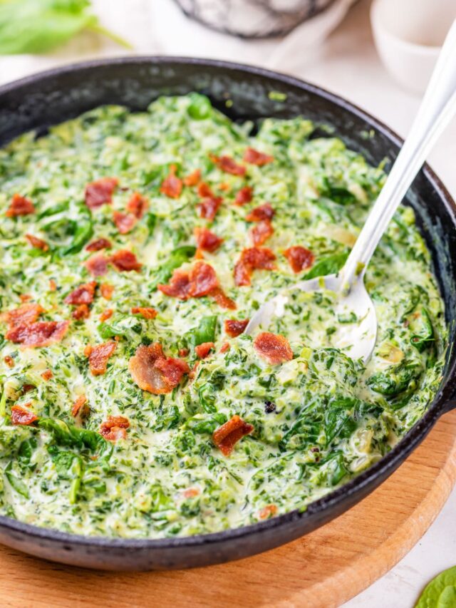 A cast iron pan filled with creamed spinach and bacon crumbles with a spoon for serving in the pan.