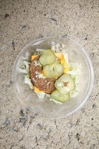 A bowl of shredded lettuce, 2 burger patties, a slice of cheese, some diced onions, 4 slices of pickles, and mustard.