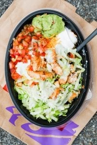 A large black bowl filled with shredded lettuce, pico de gallo, cheese, guacamole, sour cream, and chopped chicken.