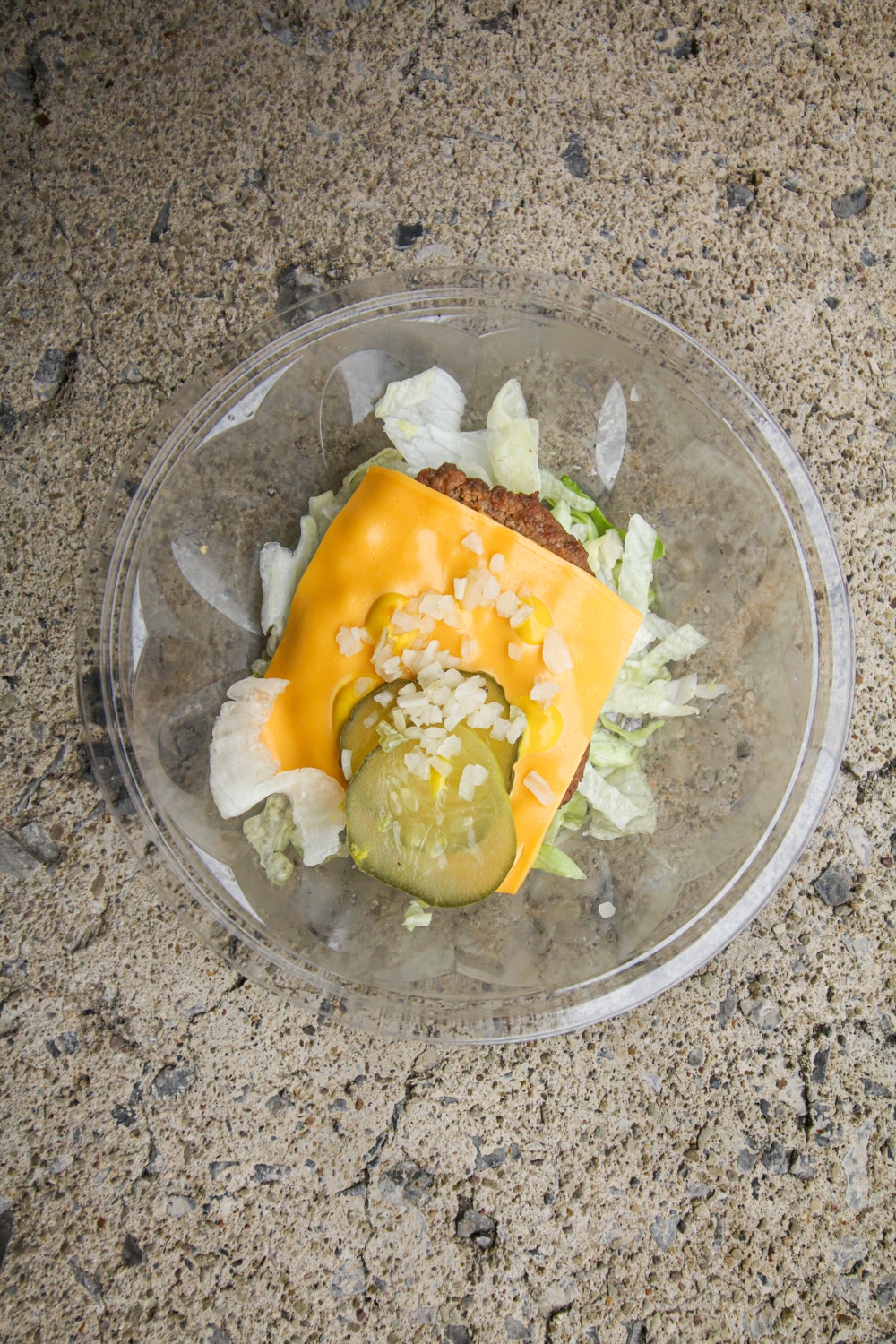 A burger patty on top of shredded lettuce and topped with a slice of cheese, mustard, pickles, and diced onion.