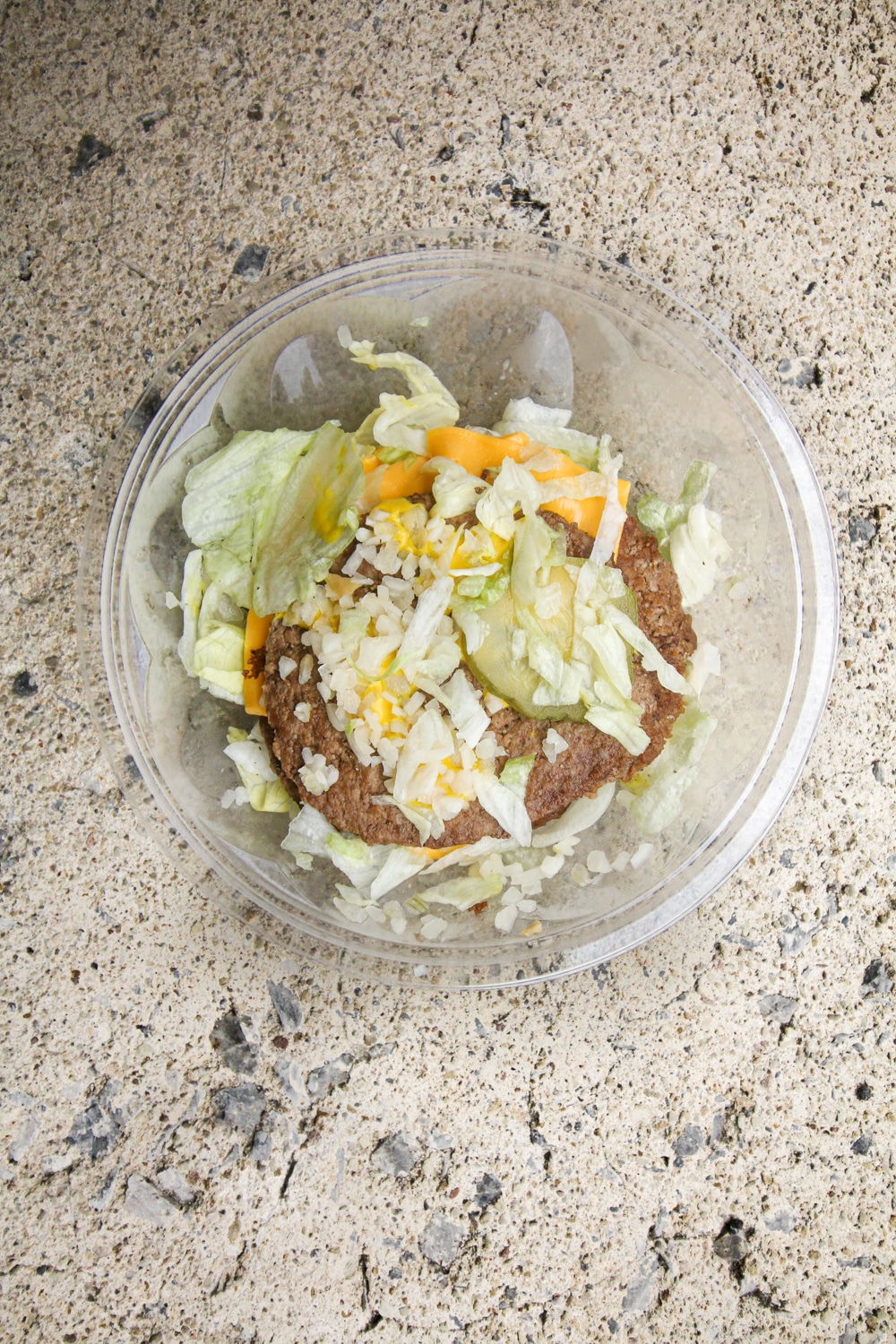 3 Burger patties in a bowl of shredded lettuce, 2 slices of cheese, sliced pickles, onions, and mustard.