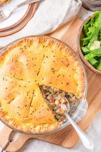 A pie plate board filled with chicken pot pie on top of a cutting and next to a bowl of lettuce.