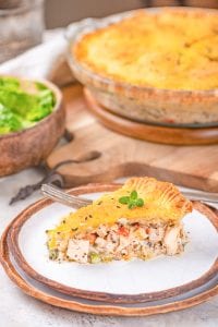 A slice of chicken pot pie on a plate with a bowl of salad in back of it, along with the rest of the pot pie.