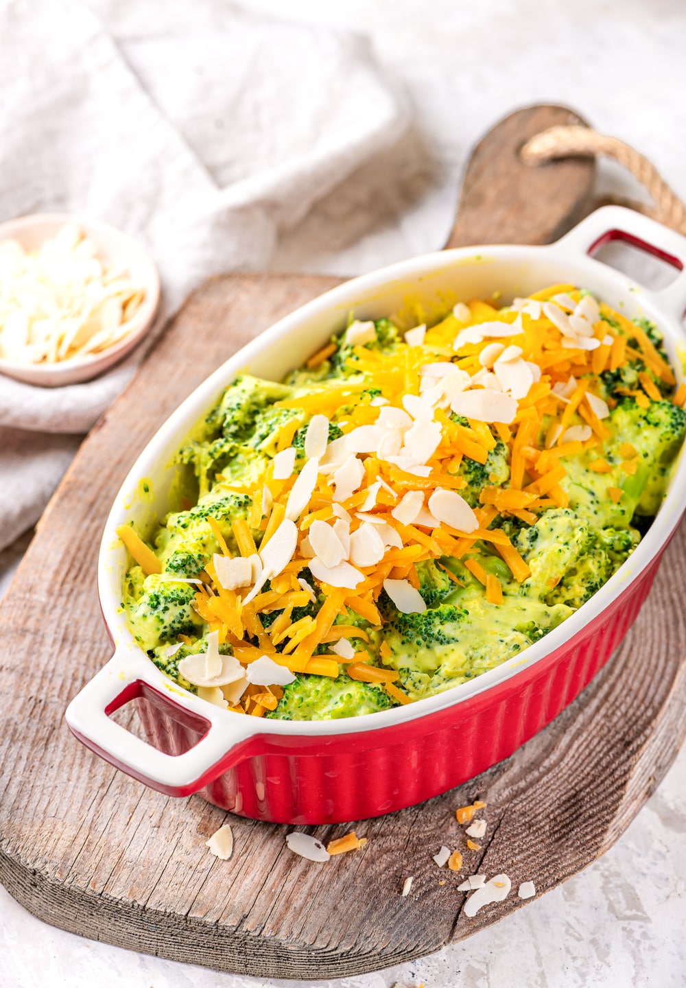A casserole dish filled with broccoli covered in cheese sauce, and topped with shredded cheddar cheese and sliced almonds.