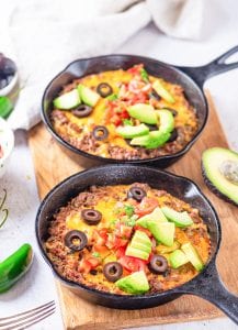 2 Cast iron pans filled with taco casserole and topped with black olives, salsa, and pieces of avocad.