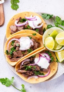 3 carne asada tacos on a plate next to a small bowl of cut up limes.