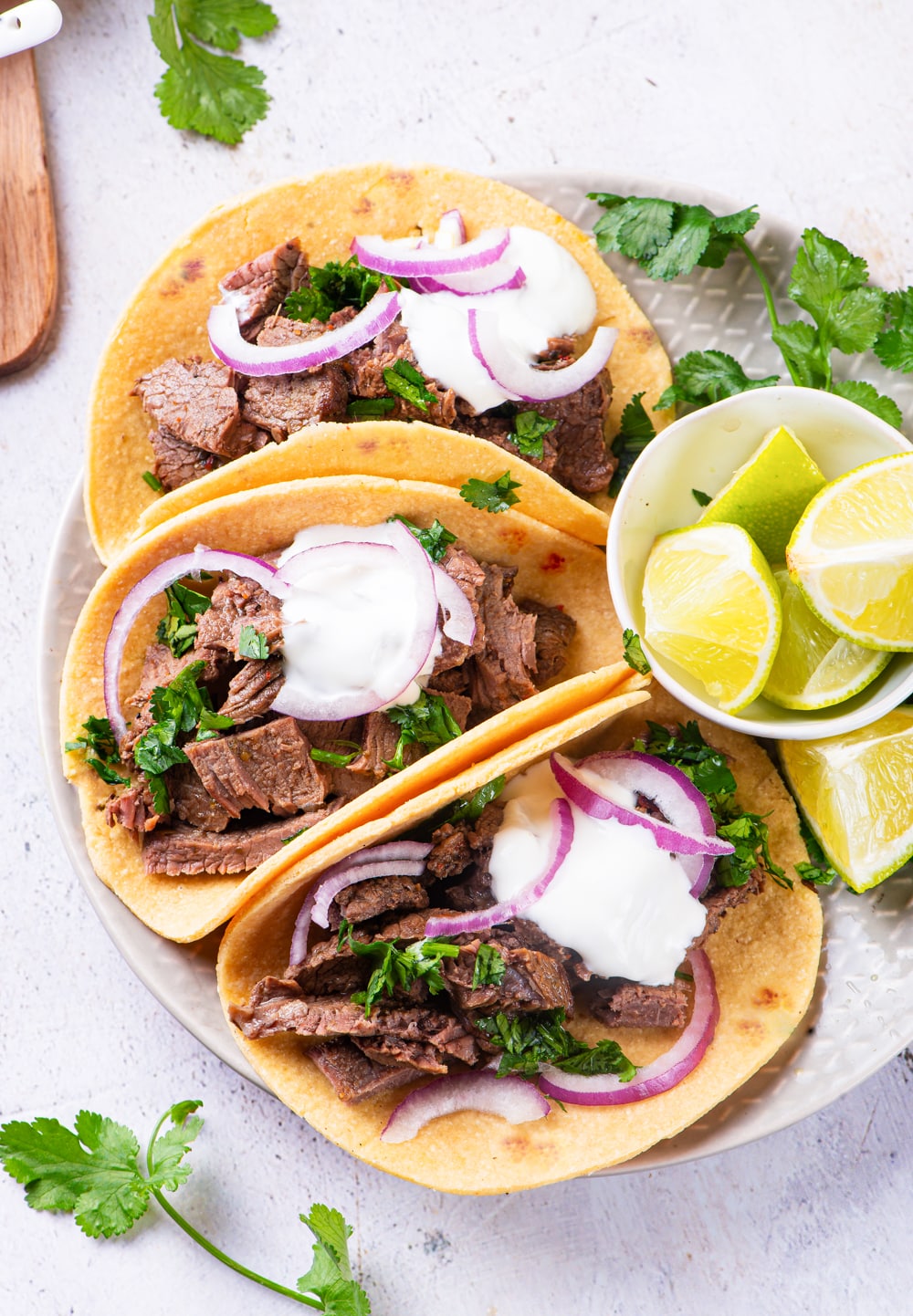 3 carne asada tacos on a plate next to a small bowl of cut up limes.