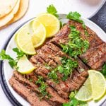 Flank steak cut up on a white plate covered in chimichurri and surrounded by lemons.