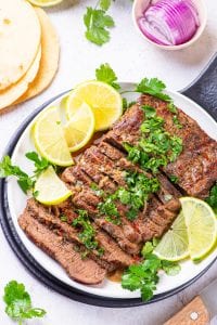 Flank steak cut up on a white plate covered in chimichurri and surrounded by lemons.