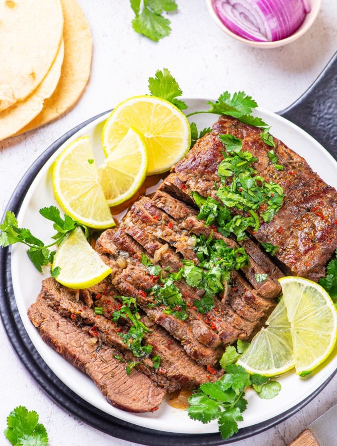 Flank steak cut up on a white plate covered in chimichurri and surrounded by lemons.