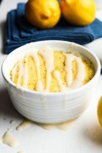 A lemon poppy seed mug cake with two lemons on a napkin behind it.