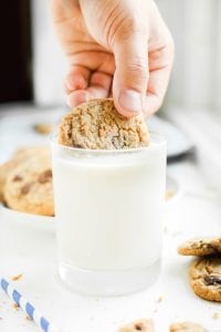 A hand dipping half of a chocolate chip cookie in a glass of milk.