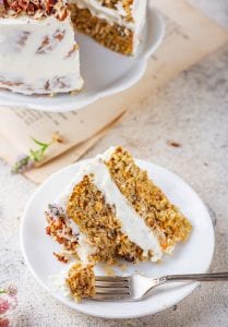 A slice of carrot cake on a white plate with a fork taking a piece from it.