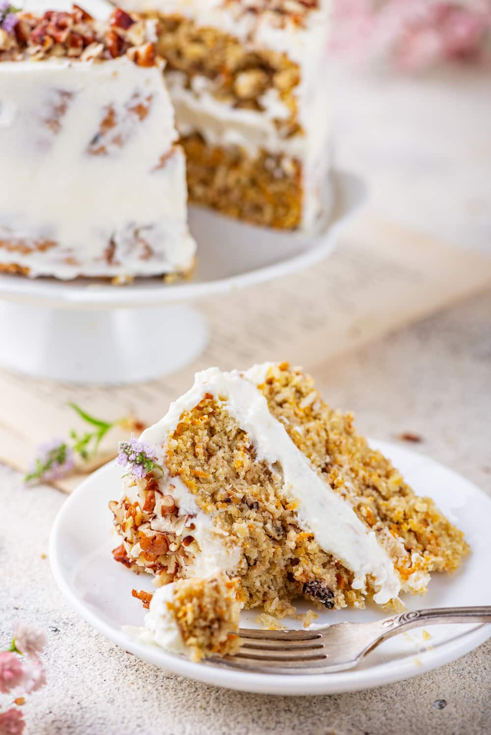 A slice of carrot cake served on a white plate, with a piece of the cake on a fork and the entire cake behind it.
