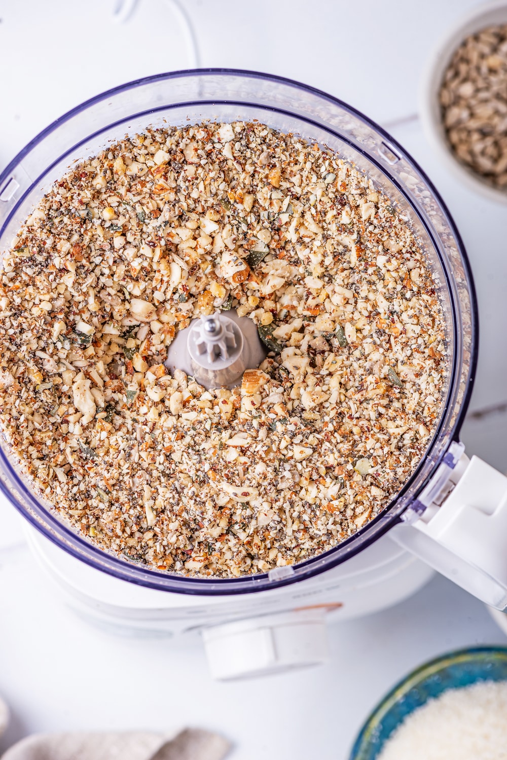 A food processor filled with crushed up nuts and seeds on a white table.