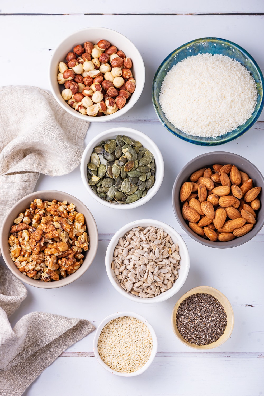 Small bowls filled with nuts, seeds, and shredded coconut on a white table.