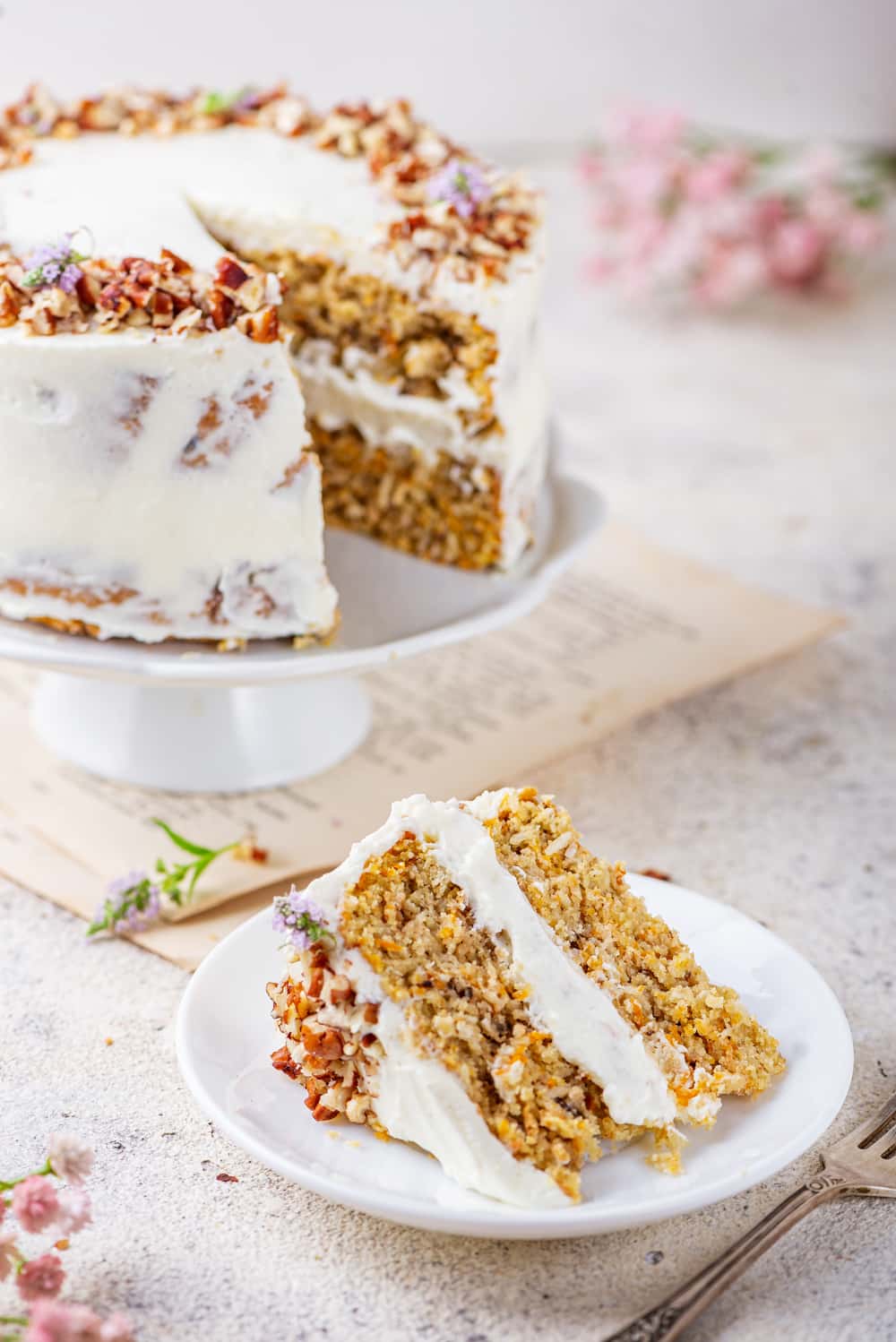 A slice of carrot cake on a white plate with the entire cake behind it.