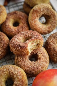 A bunch of donuts next to one another with an apple next to them on a wire rack.