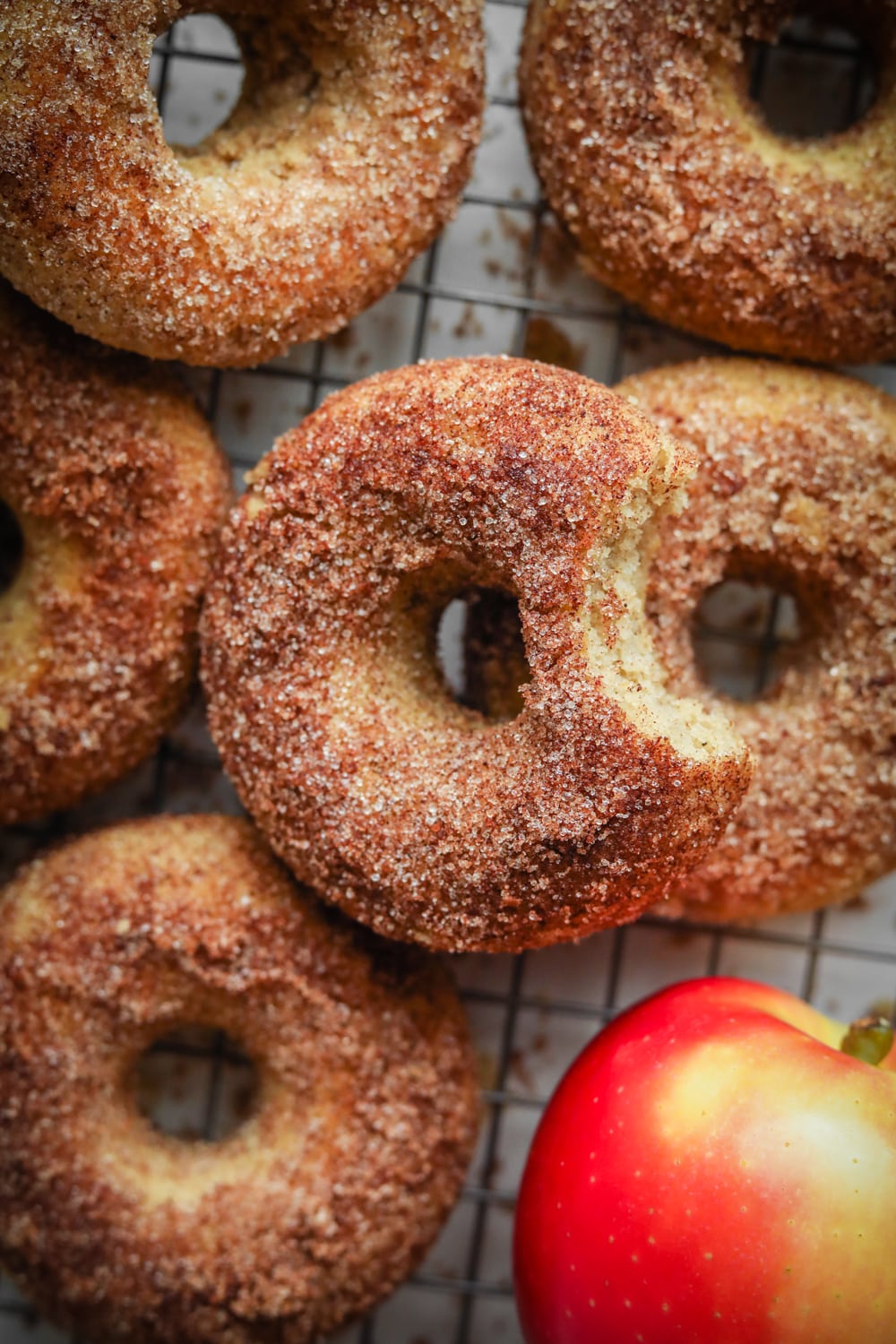 A half dozen donuts on a black wire rack next to an apple.