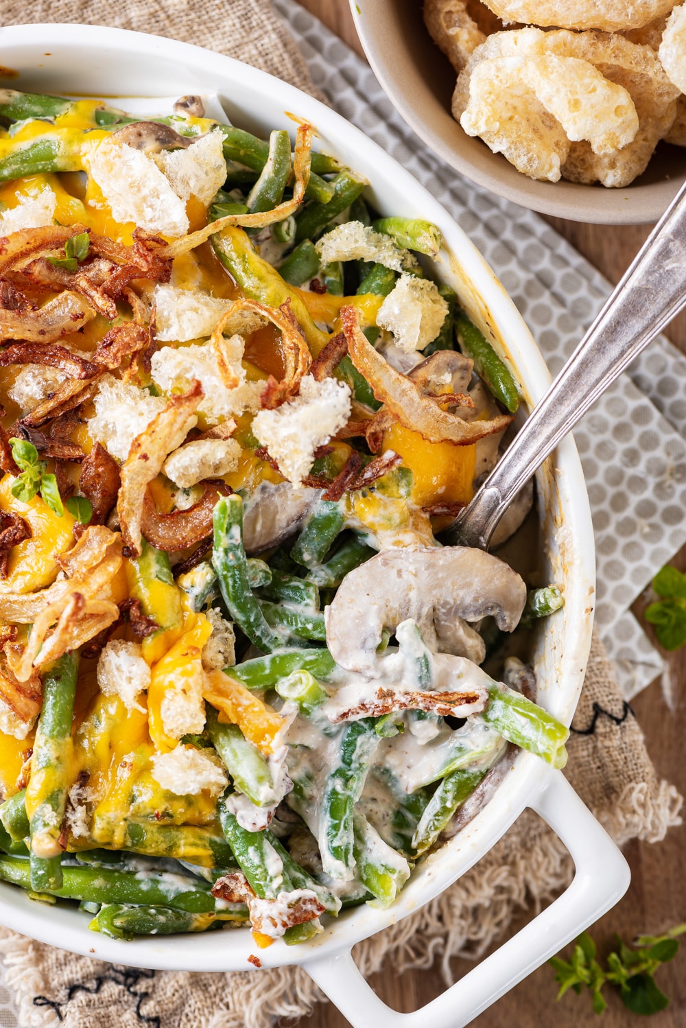 A mixture of green beans, creamy mushrooms, melted cheese, crispy onions, and pork rinds in a white casserole dish, and a fork placed in the dish as well.